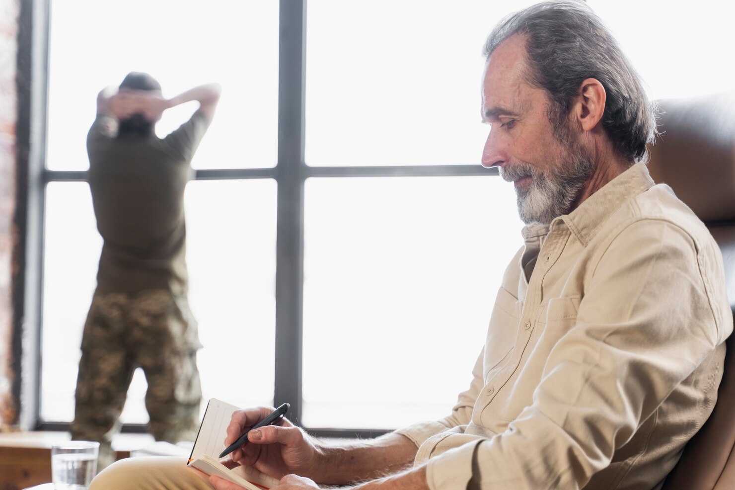 Hombre mayor escribiendo en un cuaderno mientras observa a un militar de pie junto a una ventana. La imagen muestra un ambiente reflexivo y de concentración.
