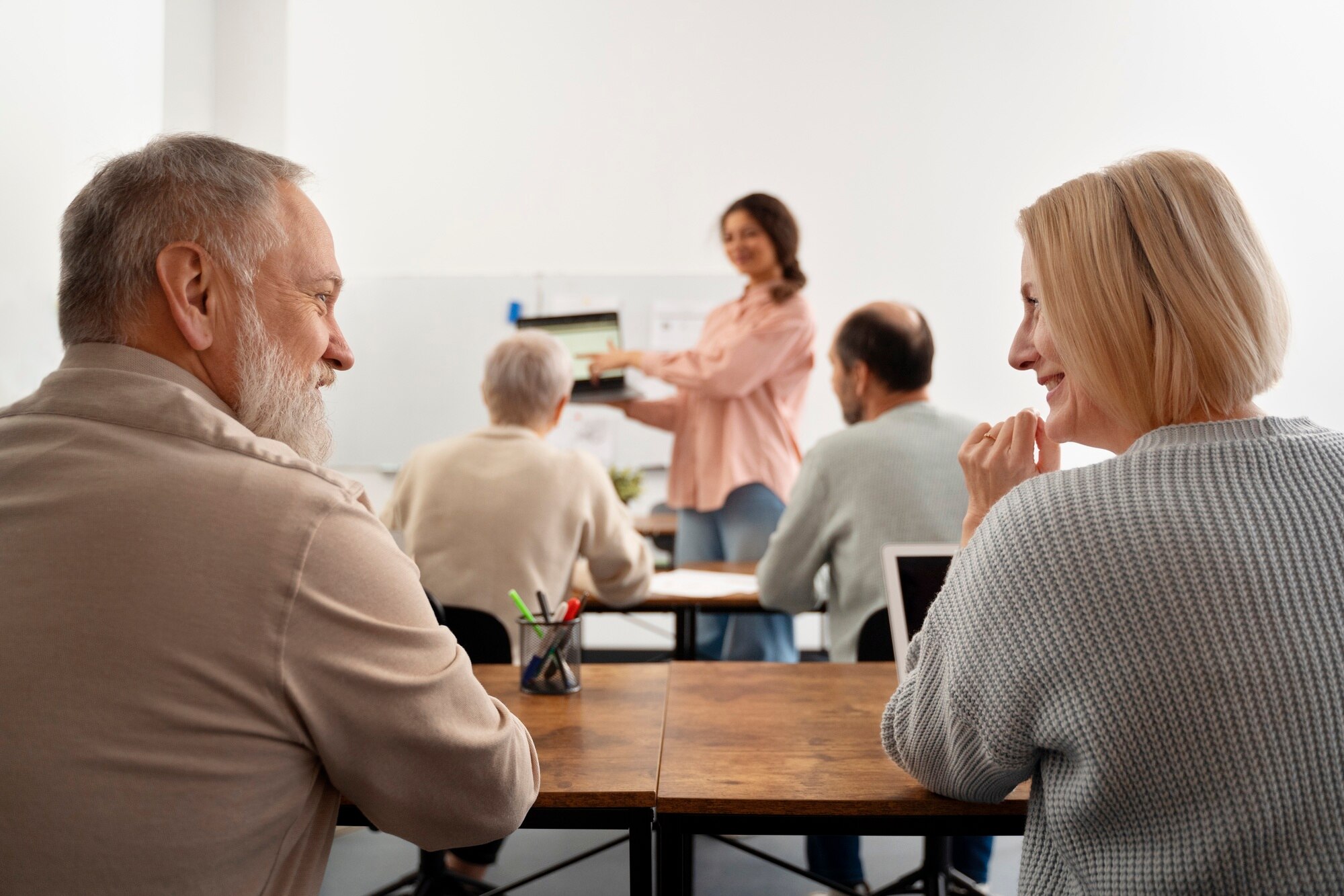 Grupo de personas mayores en una clase, escuchando a una mujer que presenta información en una computadora portátil. Se observa un ambiente de aprendizaje colaborativo y participación activa.