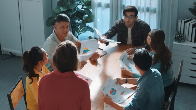 Grupo de profesionales colaborando en una reunión, analizando gráficos y documentos en una mesa. Se observan siete personas, con diversos materiales de trabajo y un ambiente de oficina iluminado.